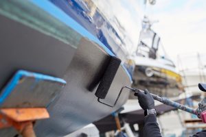 Person painting a boat's bottom with a fresh paint, using roller and black paint, outdoors in the boatyard